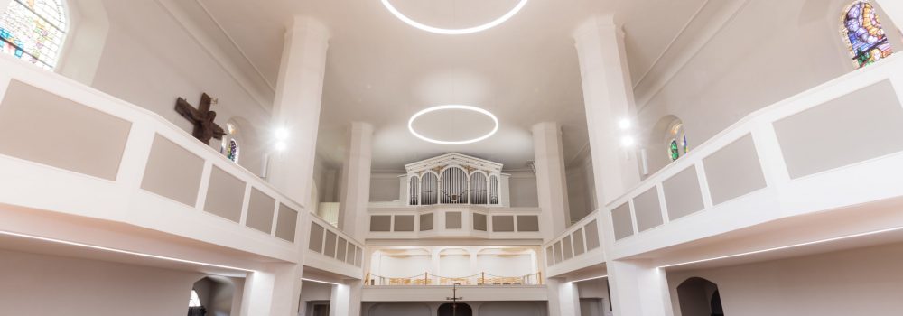 Blick über die Kirchenbänke in den Saal des Hauptschiffs - mit Ringleuchten an der Decke der Kirche