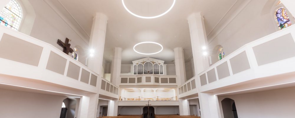 Blick über die Kirchenbänke in den Saal des Hauptschiffs - mit Ringleuchten an der Decke der Kirche